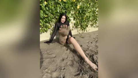 RitaMoonllight sits on the sand against a green leafy wall, wearing a black mesh bodysuit and showing her sandy bare foot close to the camera.