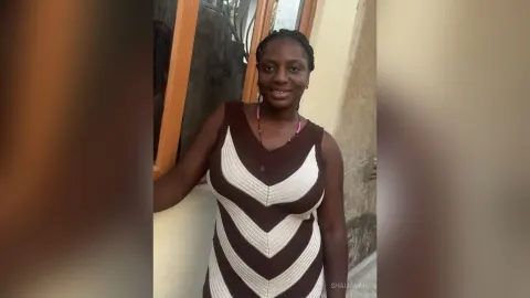 Shaluwah — Shaluwah smiles while wearing a brown and white striped sleeveless dress standing outdoors by a window.