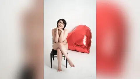 Young woman in black lingerie sitting on a chair next to a large red heart pillow in a bright studio setting.