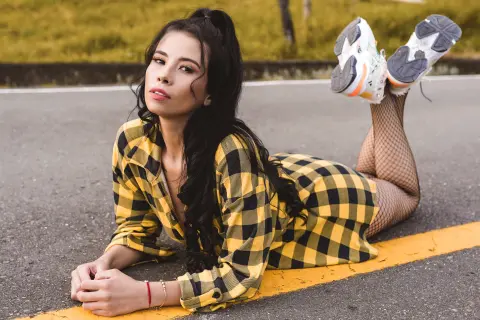 Young woman in yellow plaid outfit and fishnet tights lying on an asphalt road with a relaxed mood.