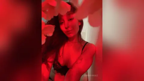 SammyRouz poses in a black lace bra with her long dark hair, framed by red flowers in a soft-lit setting.