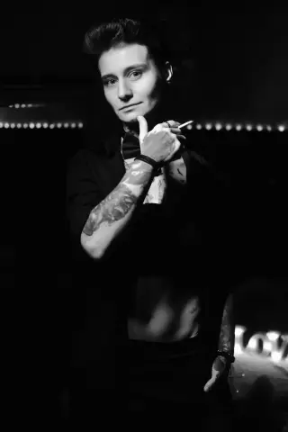 Moody black and white portrait of a tattooed man in a dark shirt holding a cigarette in dim lighting.