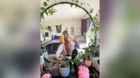 Young woman in an orange sweater and white striped tights taking a mirror selfie in a cozy, plant-filled bedroom.