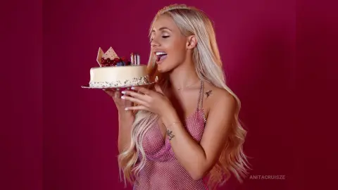 Anita smiles while holding a small white cake topped with berries and chocolate, wearing a pink polka-dot slip dress.