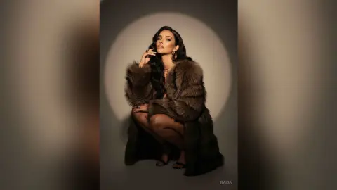Elegant woman in a luxurious brown faux fur coat posing against a dramatic spotlight background.