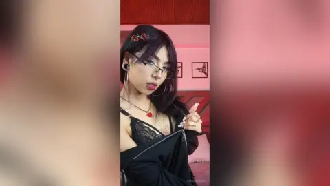 Young woman with glasses and dark hair wearing a black lace top in a soft pink aesthetic room.