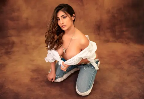 Beautiful woman in a white off-the-shoulder shirt and denim jeans posing against a warm brown studio background.