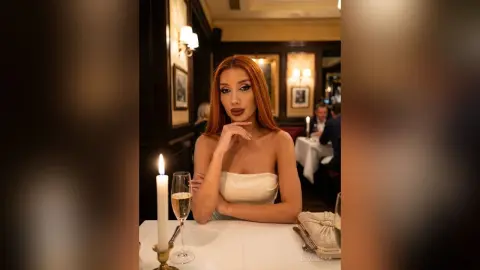 DeviNessa poses at a white table in a white strapless top, looking at the camera with long red hair and a lit candle nearby.