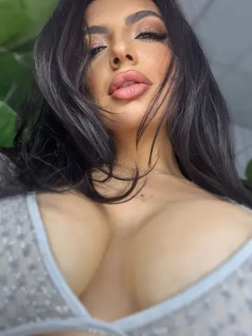NoraSin poses close-up, showing her long dark hair and large breasts in a sparkly silver bra or top.