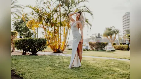 Elegant woman in a white slit maxi dress posing on a sunny tropical lawn near palm trees.