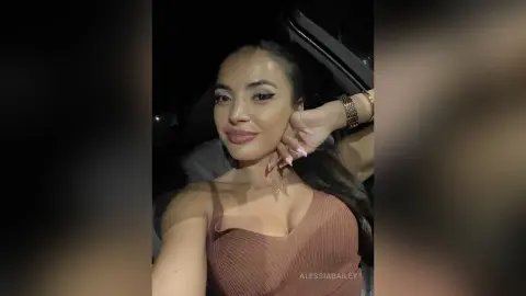Smiling woman in a brown ribbed tank top sitting in a car at night with a glamorous makeup look.