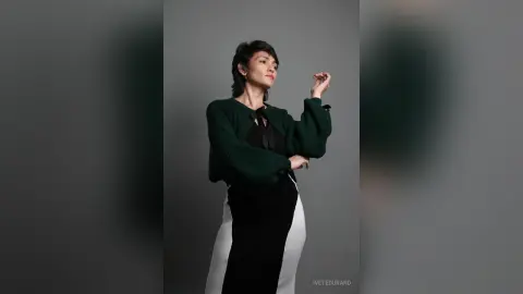 IveteDurand poses in a dark green long-sleeve top and a black and white skirt against a plain grey background.
