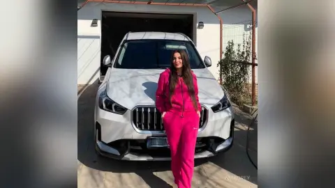 Stylish woman in a pink tracksuit standing in front of a white luxury SUV in a sunny driveway.