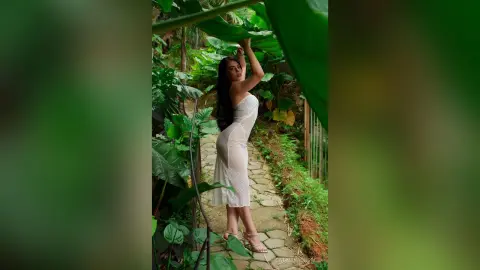 Elegant woman in a white lace dress posing amidst lush tropical greenery in a serene garden setting.