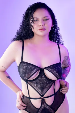 Young woman with curly hair wearing black lace lingerie against a soft purple studio background.