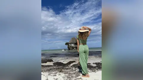 Woman in green patterned jumpsuit and sun hat standing on a tropical beach facing a seaside hut under blue skies.