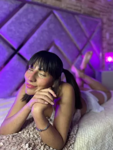 Smiling woman with dark bangs in white lingerie relaxing on a bed under soft purple mood lighting.