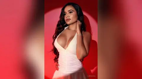 LaraCole poses in a low-cut white dress with her long dark hair down against a red background.