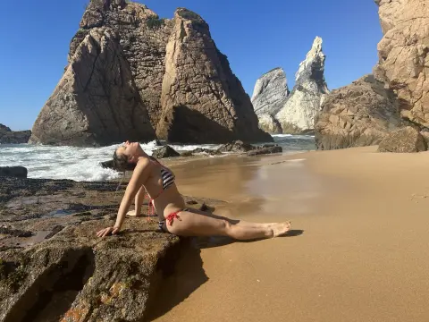 MagneticAmy poses naked on a beach rock, arching her back and looking up toward the sky in a striped bikini.