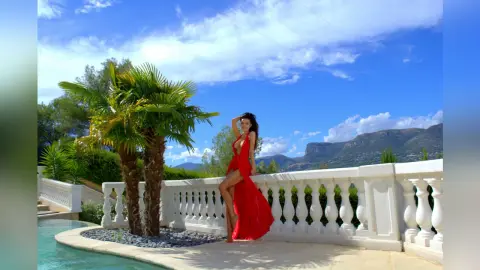 Elegant woman in a long red gown posing by a luxury villa pool under a bright blue sky.