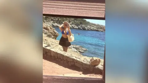 Young woman in a blue bikini top and black skirt enjoying a sunny summer day by the rocky Mediterranean coast.