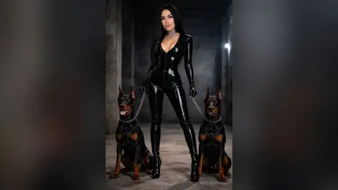 Elegant woman in a black latex bodysuit posing with two Doberman Pinschers in a moody, dark studio setting.