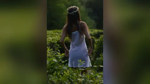 Dreamy woman in a white summer dress and flower crown walking through a lush green field at sunset.