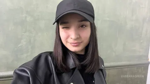 Smiling young woman in a black leather jacket and baseball cap winking against a textured grey wall.