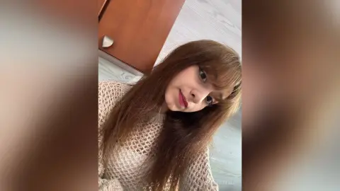 Young woman with long brown hair wearing a white crochet top in a bright indoor setting.