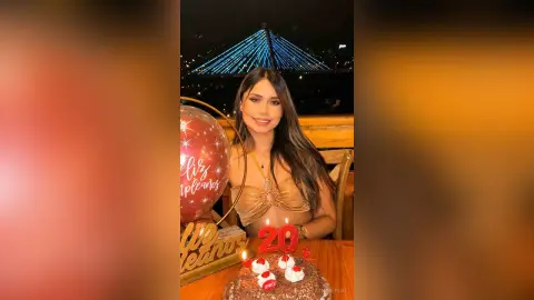 Young woman in a gold bikini top celebrating her 20th birthday with a chocolate cake and red balloons at night.