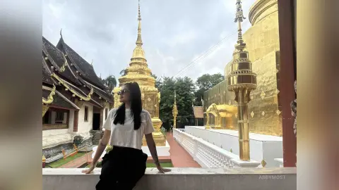 Young woman in a white t-shirt sitting by a golden Buddhist temple under a cloudy sky.
