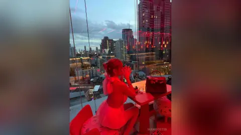 Stylish woman in a red outfit sitting on a rooftop balcony overlooking a city skyline at twilight.