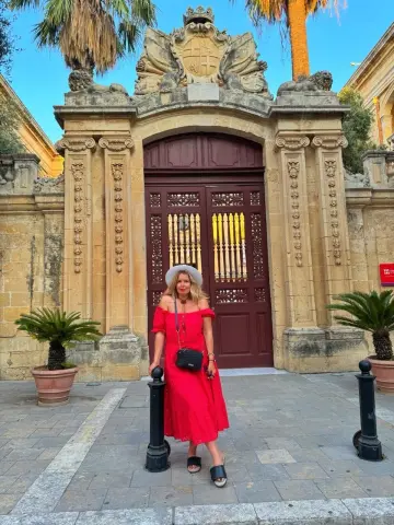 NikaZen stands in front of a large ornate stone gate wearing a red off-the-shoulder dress and a white sun hat.