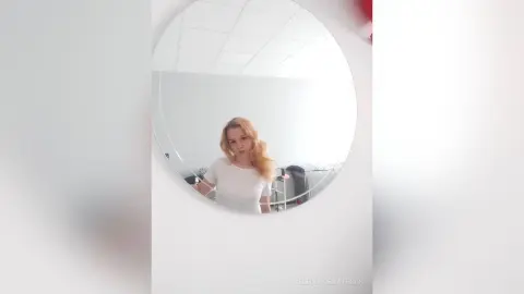 Blonde woman in a white t-shirt reflected in a round mirror within a bright, minimalist room.