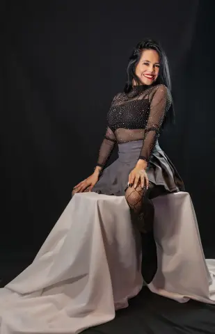 Smiling woman in a black sheer sequin top and grey skirt sitting on a draped white pedestal against a dark backdrop.