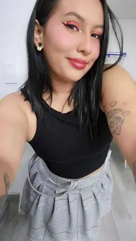 Smiling woman with dark hair and winged eyeliner wearing a black tank top and grey plaid skirt indoors.