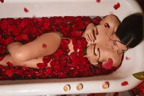 Nude woman relaxing in a white bathtub filled with red rose petals for a romantic and sensual spa mood.
