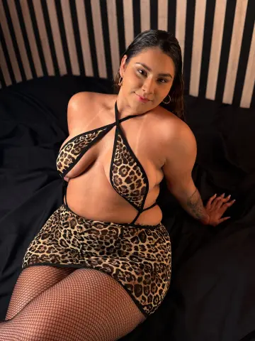 Curvy woman in leopard print lingerie and fishnet tights relaxing on a black bed against a striped wall.
