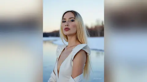 Blonde woman in an elegant white dress posing by a serene frozen lake during a peaceful winter sunset.