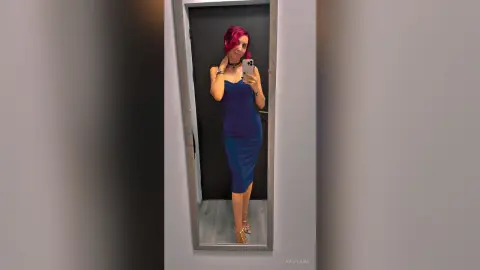 Woman with red hair in a blue midi dress taking a mirror selfie in a modern hallway.