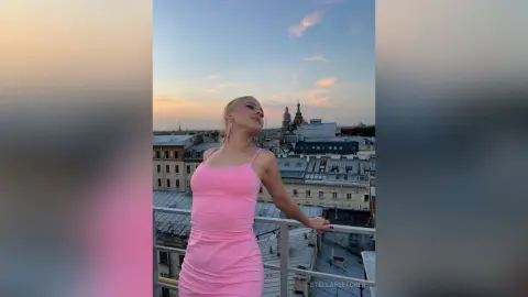 StellaFletcher stands on a balcony overlooking a city skyline, wearing a tight pink tank top and matching pink skirt.