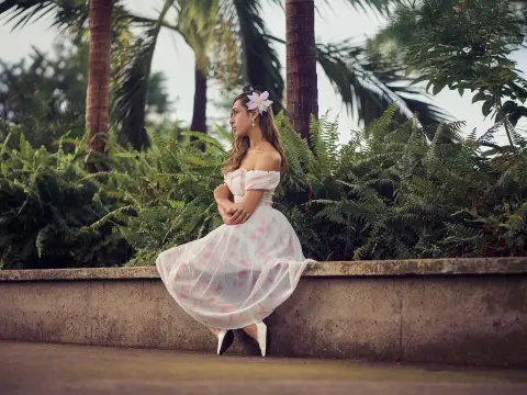 Woman in a white floral summer dress sitting on a stone ledge in a lush tropical garden setting.