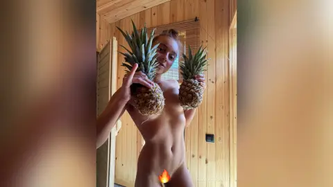 Young woman holding two pineapples in a rustic wooden cabin setting with a natural, summer vibe.