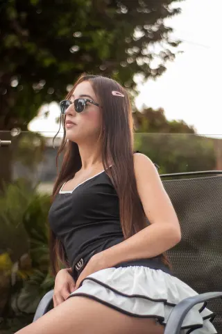 AlaskaWilliamss sits outdoors in a black tank top and white skirt, wearing sunglasses and looking away.