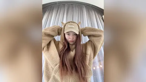 Young woman in a cozy tan fleece hoodie and matching cap posing indoors against sheer white curtains.
