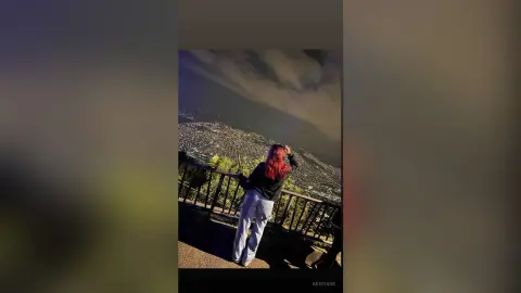 Red-haired woman in a black top and light jeans overlooking a city landscape at dusk.