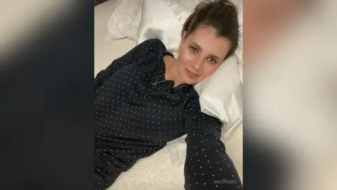 Smiling woman in navy polka dot pajamas relaxing on a white bed in a cozy bedroom setting.