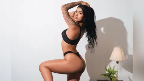 Tattooed woman in black bikini posing against a white wall in a bright, minimalist studio setting.