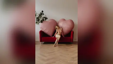 Blonde woman in a red bikini sitting on a red sofa against large pink heart pillows in a minimalist room.