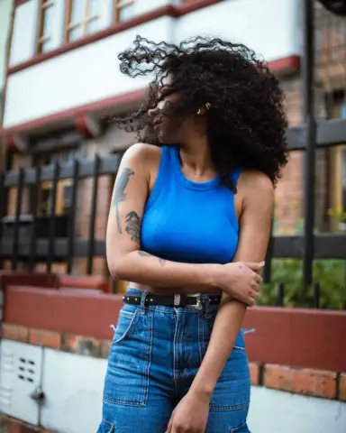 Young woman with curly hair in a blue crop top and denim jeans posing outdoors in a city setting.